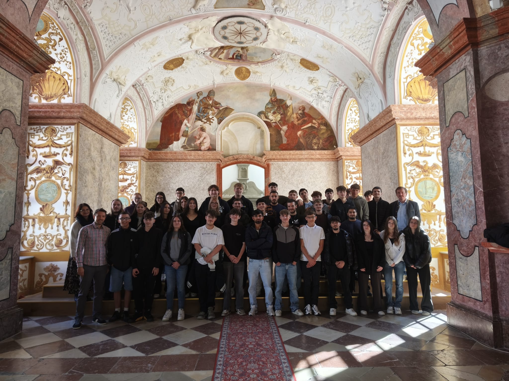 Gruppe von Schülern und Lehrern vor prunkvoller Deckenmalerei in einem historischen Gebäude